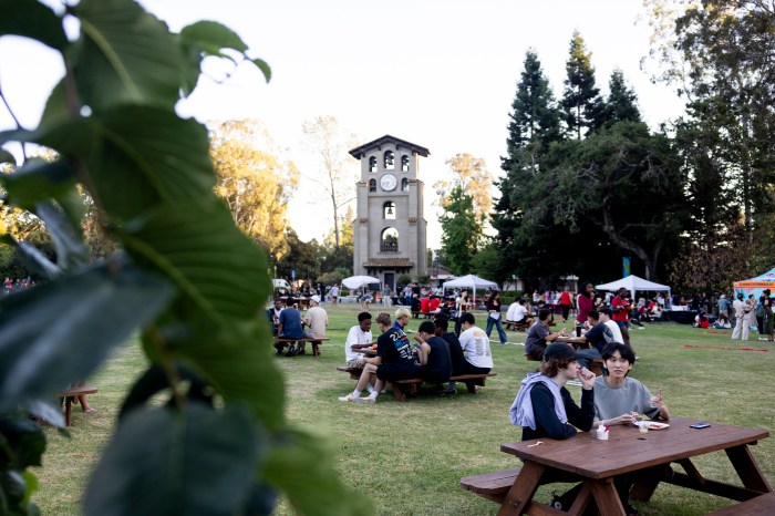 Students sit at picnic tables on a lawn eating from food trucks, with a bell tower and tented vendor stalls in the background, viewed through foreground leaves.