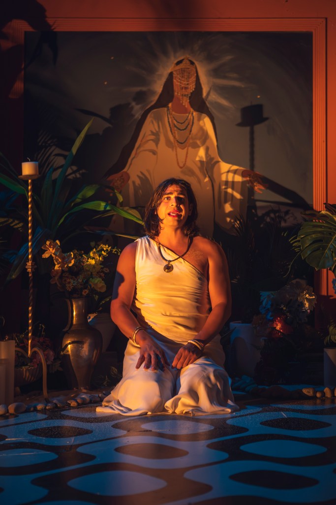 kneeling person in white dress with goddess painting and plants behind him 