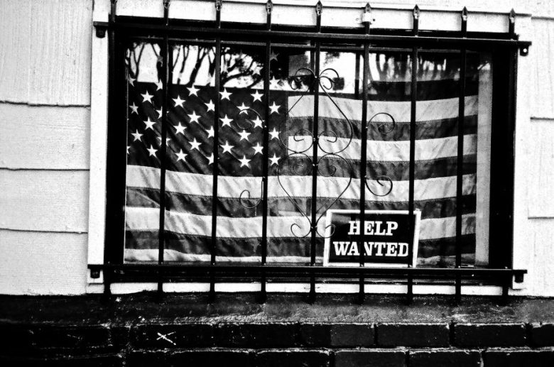 A barred window displays an American flag with a “Help Wanted” sign placed in front of it.
