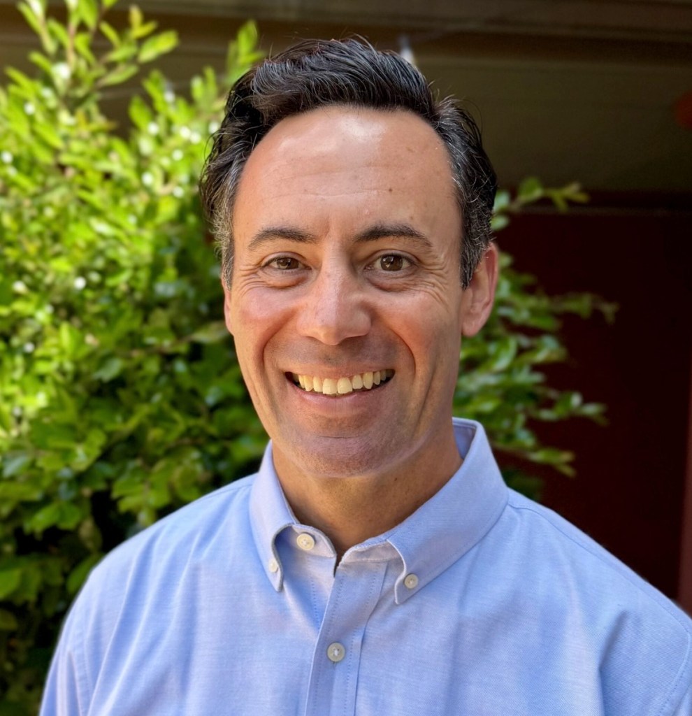 Headshot of a man with dark hair, a light blue shirt, and a wide smile, in front of green foliage.