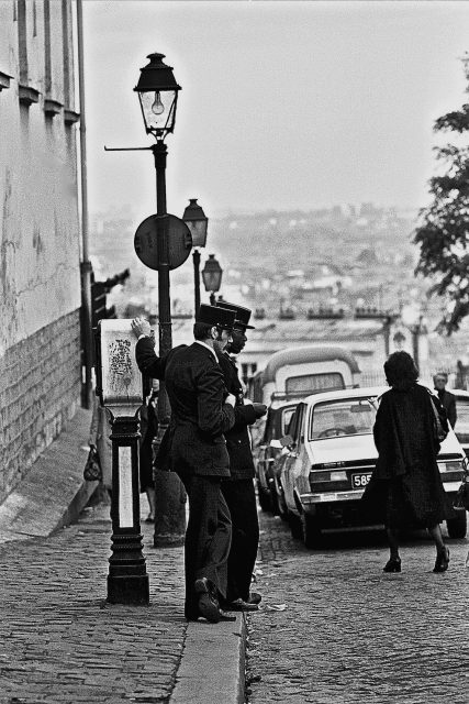 Two men in uniforms stand by a streetlamp on a cobblestone hill, with parked cars and a woman walking away in the background.
