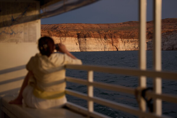A person takes a picture during a tour on Lake Powell at the Glen Canyon National Recreation Area, Wednesday, July 15, 2026, near Page, Ariz. A bathtub ring of light minerals on the canyon wall shows the high water mark of the reservoir. (AP Photo/John Locher, File)