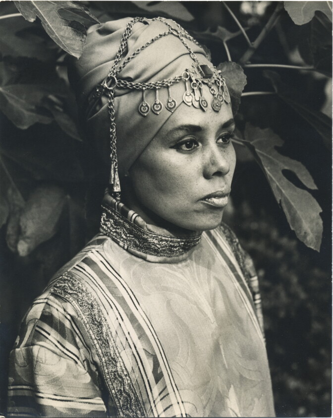 A female presenting person wears a turbn and robe with metal jewelry.