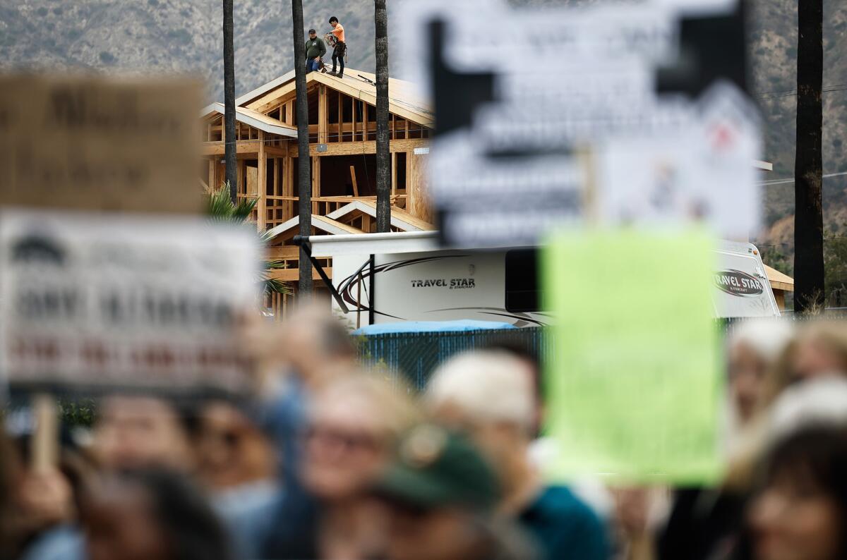 Protesters holding signs while, in the distance, construction workers stand atop the wooden frame of a house