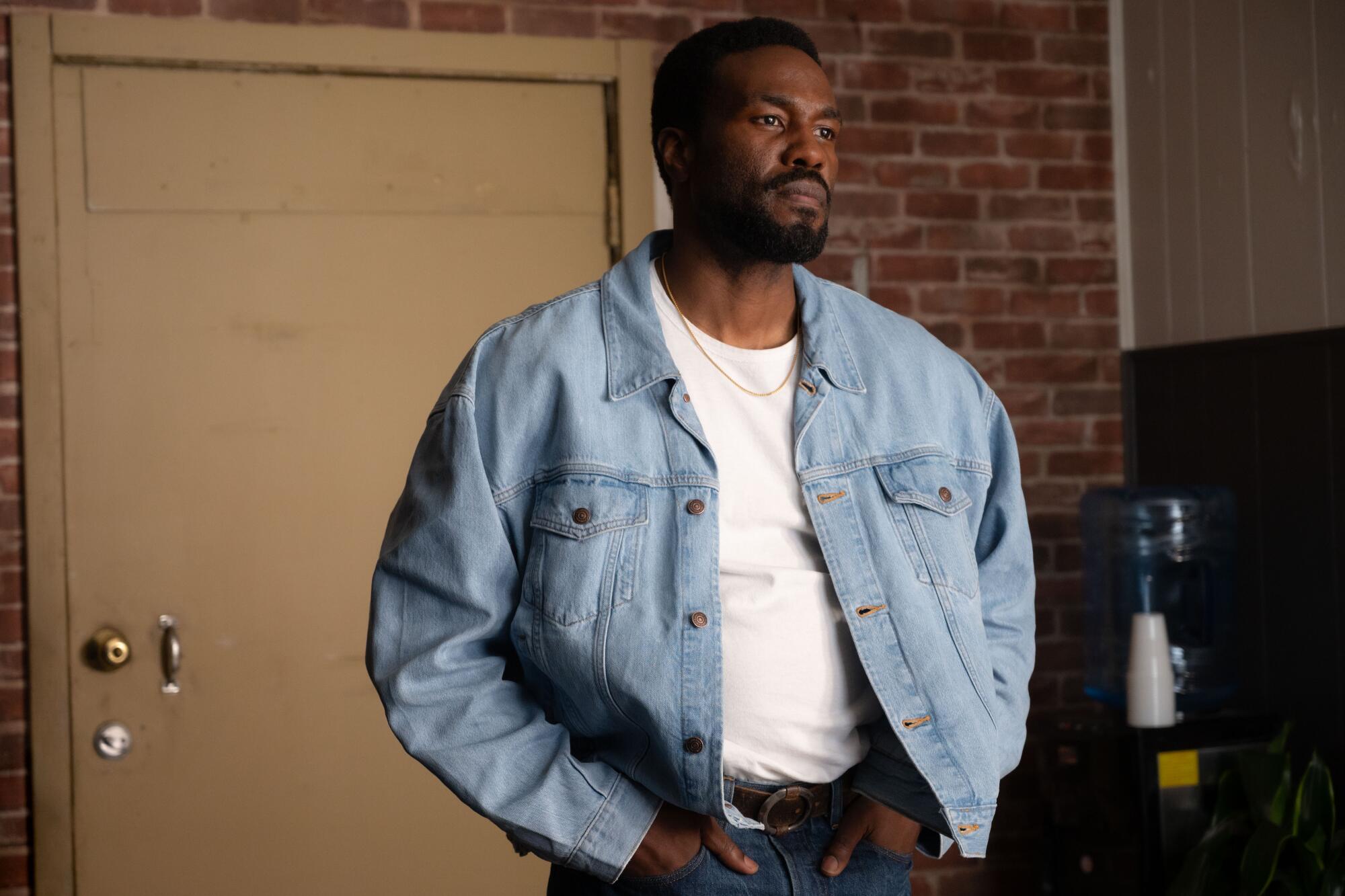 A man in a denim jacket and white T-shirt stands with his hands in his pockets.