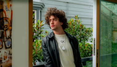 A young man with curly hair wears a black leather jacket and white shirt, standing near a glass door with greenery and a white house exterior in the background.