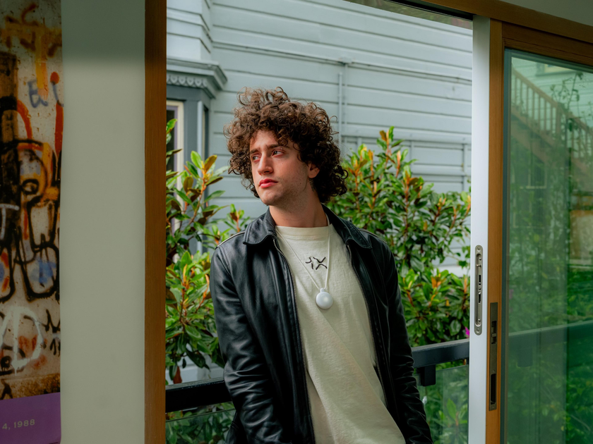 A young man with curly hair wears a black leather jacket and white shirt, standing near a glass door with greenery and a white house exterior in the background.
