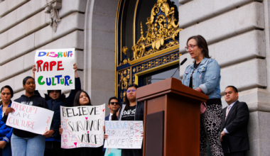 A woman speaks at a podium outside a grand building while people behind her hold signs saying "Disrupt Rape Culture" and "Believe Survivor Voices."