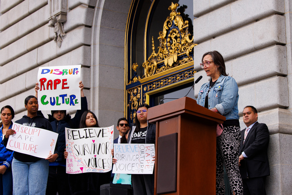 A woman speaks at a podium outside a grand building while people behind her hold signs saying "Disrupt Rape Culture" and "Believe Survivor Voices."