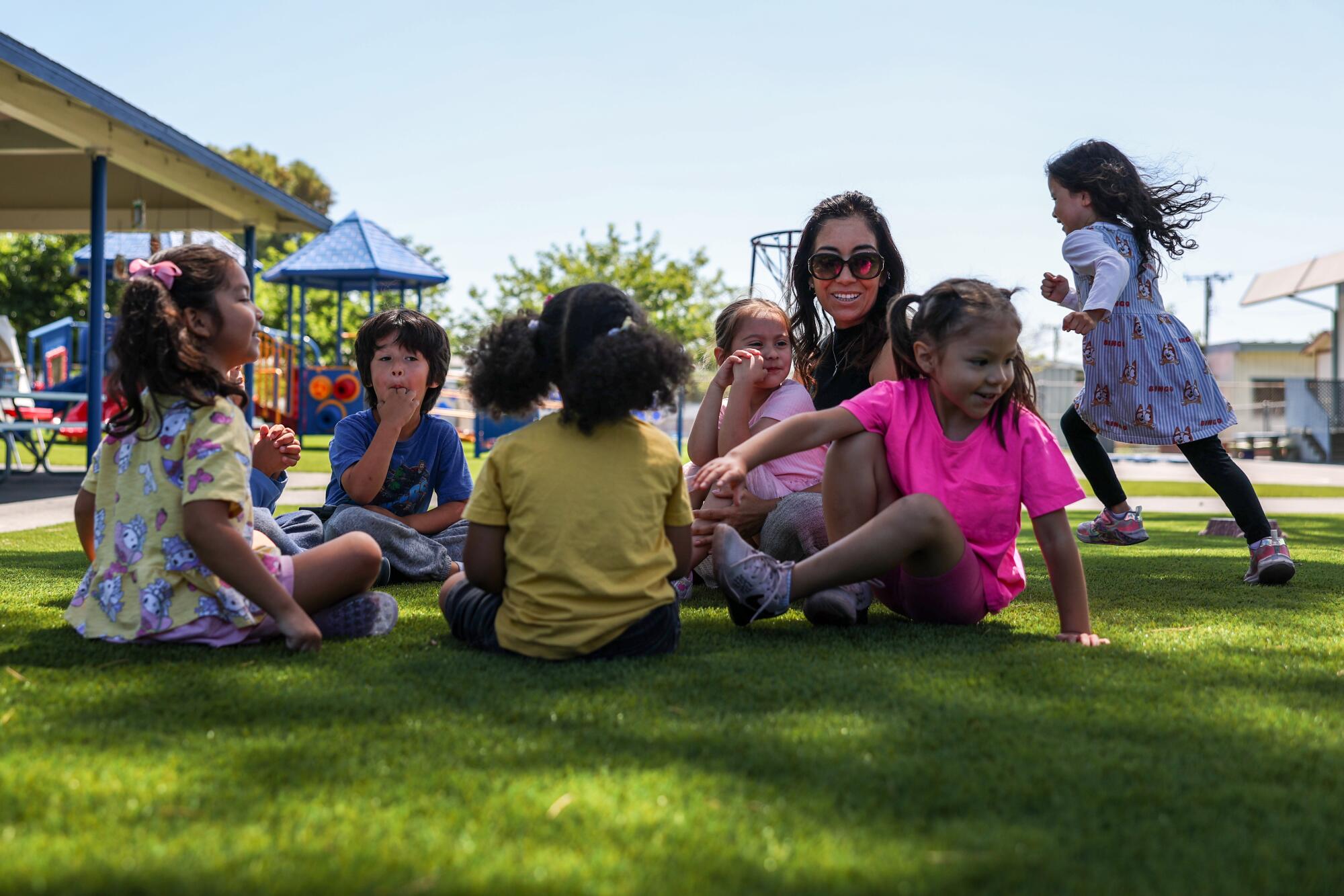 A teacher sits in a circle on the grass with six preschoolers as one runs around them in a game of 'Duck, Duck, Goose'