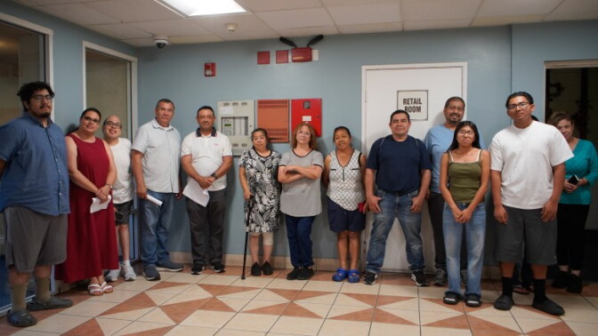 A group of people look forward for a photo while standing in a building lobby. Some are holding pieces of paper at their sides under a bright fluorescent light.
