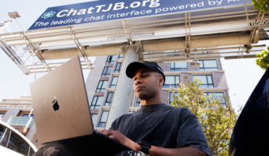 A man wearing a black cap and dark shirt uses a laptop outdoors under a large ChatTJB.org billboard on a sunny day next to a building and trees.