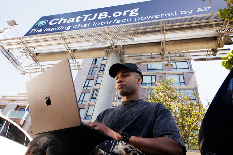 A man wearing a black cap and dark shirt uses a laptop outdoors under a large ChatTJB.org billboard on a sunny day next to a building and trees.