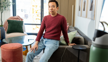 A man in a red sweater and light blue jeans sits on the edge of a gray sofa in a well-lit living room with framed pictures and colorful cushions.