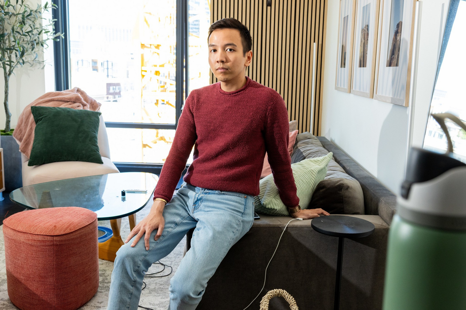 A man in a red sweater and light blue jeans sits on the edge of a gray sofa in a well-lit living room with framed pictures and colorful cushions.