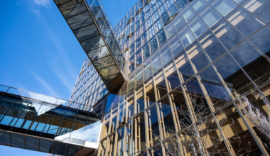 Glass buildings with reflective windows are connected by transparent sky bridges against a clear blue sky, with bare tree branches visible below.