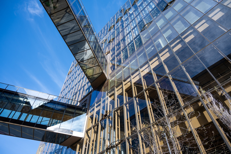 Glass buildings with reflective windows are connected by transparent sky bridges against a clear blue sky, with bare tree branches visible below.