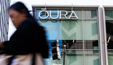 A large "OURA" sign is mounted on a glass building, with a blurred person walking past in the foreground and a traffic light reflected in the window.