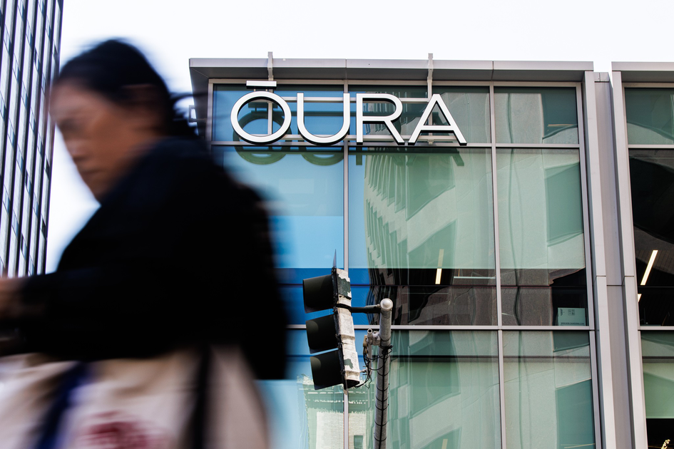 A large "OURA" sign is mounted on a glass building, with a blurred person walking past in the foreground and a traffic light reflected in the window.