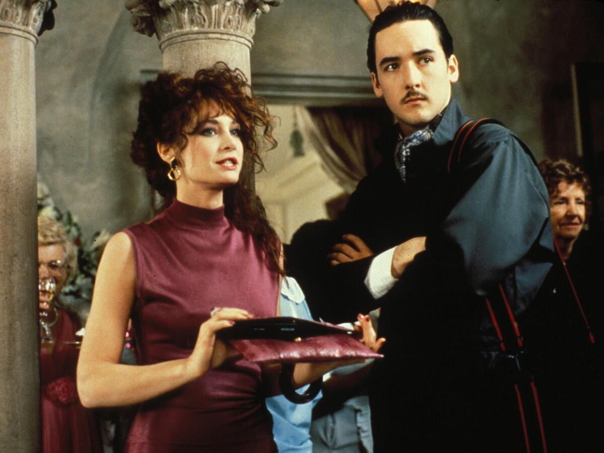 A woman in a burgundy dress stands with an intense-looking man.