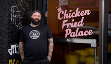 A bearded man with tattoos and a black beanie stands smiling by a restaurant door with the words "Chicken Fried Palace" in pink.