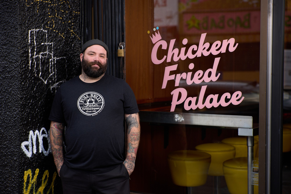 A bearded man with tattoos and a black beanie stands smiling by a restaurant door with the words "Chicken Fried Palace" in pink.