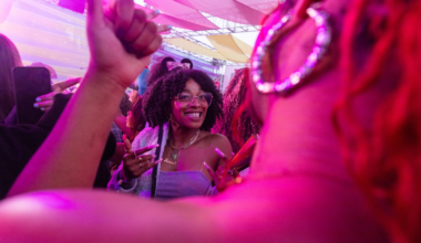 A smiling woman with curly hair and glasses wears cat ears and a necklace, surrounded by people under a pink-lit canopy.