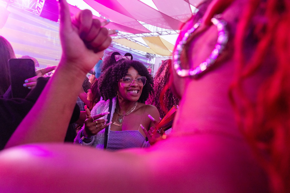 A smiling woman with curly hair and glasses wears cat ears and a necklace, surrounded by people under a pink-lit canopy.