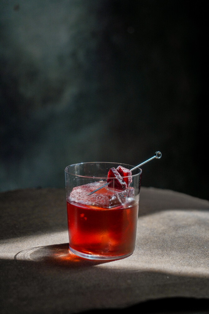 Close-up of a deep red vermouth cocktail in a rocks glass, poured over a single large, cloudy ice cube. A thin metal cocktail pick rests across the rim, skewering two dark red pieces of dried fruit garnish. Light catches the glass, casting a warm reddish glow across the table below.