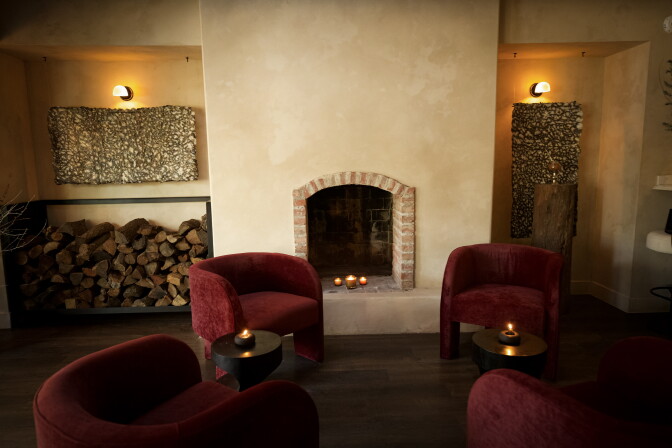 Interior shot of ALTO's lounge area at dusk, showing four burgundy velvet armchairs arranged around small black side tables with lit candles. A brick fireplace with three votive candles sits at the center of a warm, plaster-textured wall, flanked by two textured tapestry-like wall hangings under sconce lighting. A neat stack of firewood is visible to the left.
