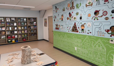 A colorful mural with children playing and various objects decorates the wall, a table with a wooden block tower and scattered blocks stands in front of a bookshelf.