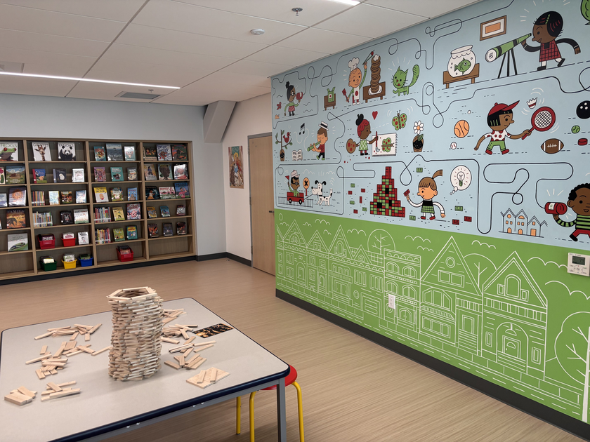 A colorful mural with children playing and various objects decorates the wall, a table with a wooden block tower and scattered blocks stands in front of a bookshelf.