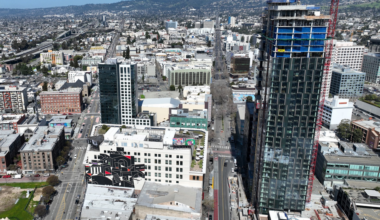 A cityscape with a tall under-construction skyscraper, surrounding mid-rise buildings, streets, and a distant hilly landscape under a clear blue sky.