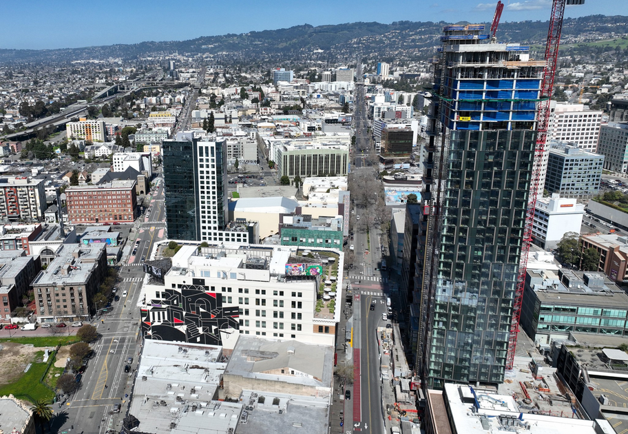 A cityscape with a tall under-construction skyscraper, surrounding mid-rise buildings, streets, and a distant hilly landscape under a clear blue sky.