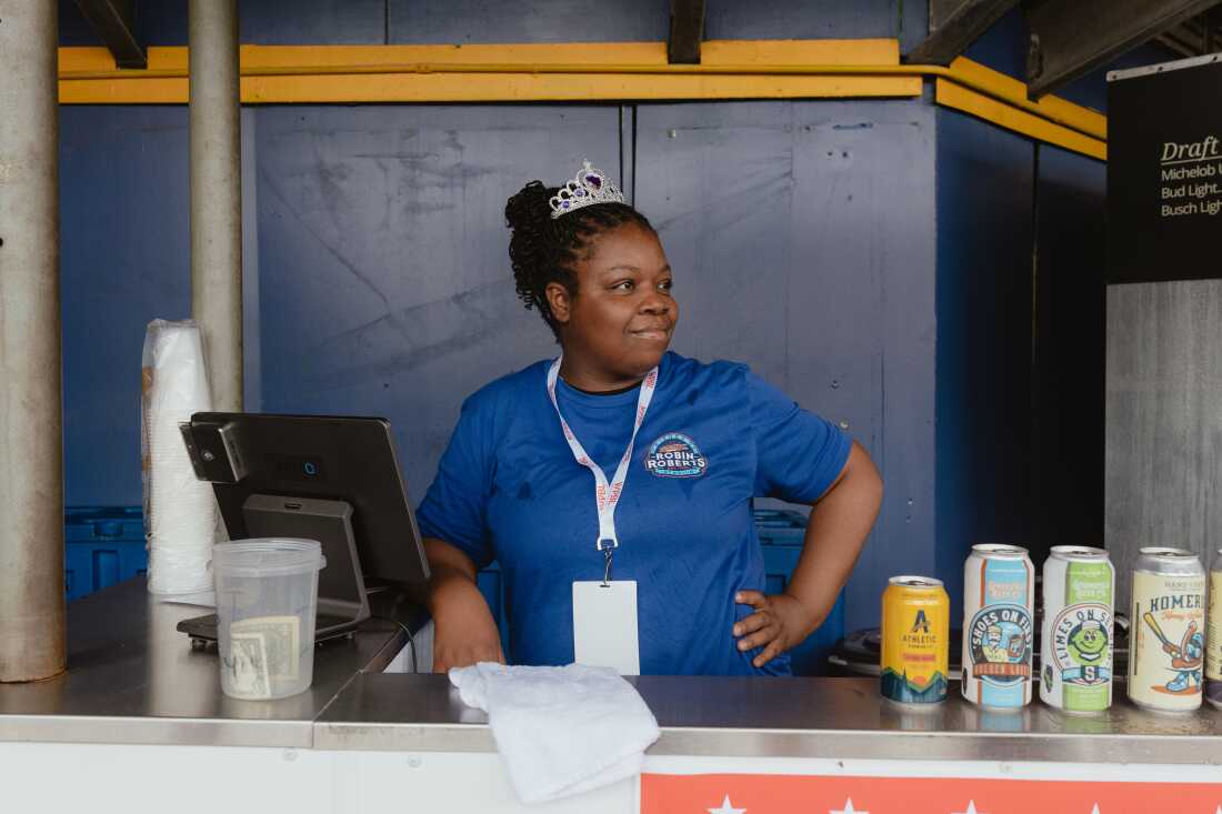 Melody, concessions worker at Robin Roberts Stadium, awaits her first customers on Opening Day of the Women's Pro Baseball League. Springfield, IL, August 1, 2026.