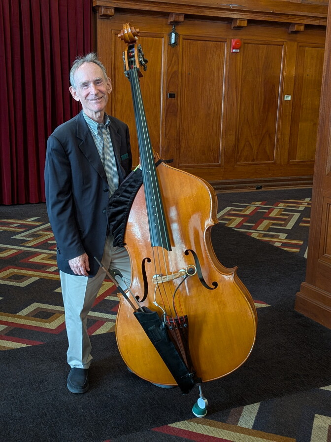 A man in a dinner jacket standing next to a huge jazz bass that's taller than him.