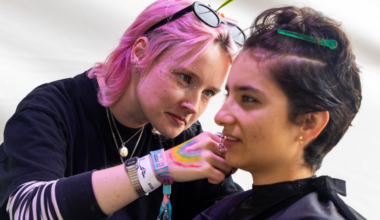 A person with pink hair and sunglasses on their head carefully applies makeup or face paint to another person with short dark hair, who wears a black cape.