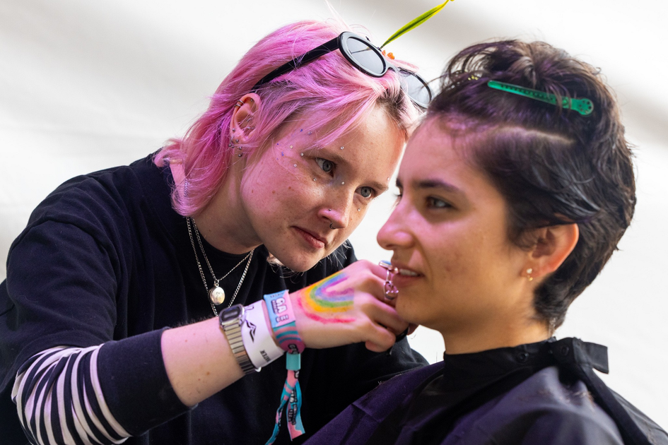 A person with pink hair and sunglasses on their head carefully applies makeup or face paint to another person with short dark hair, who wears a black cape.