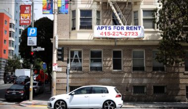 A white hatchback car is parked on the street in front of a brick building with a large "APTS FOR RENT" sign and a contact number.