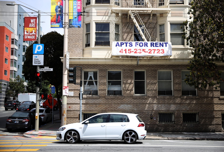 A white hatchback car is parked on the street in front of a brick building with a large "APTS FOR RENT" sign and a contact number.