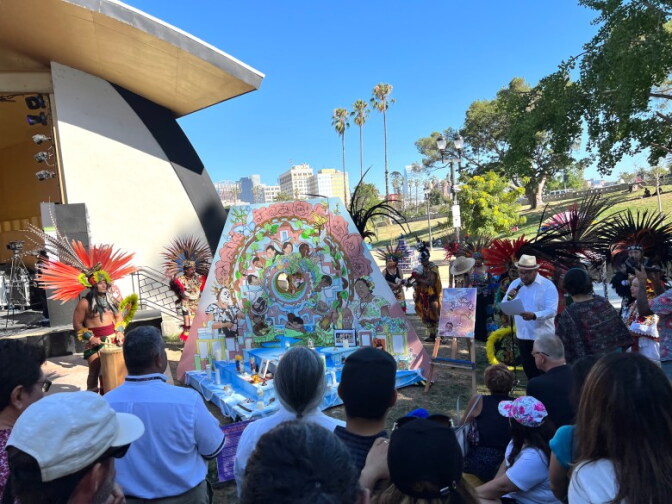 A group of people gather around a colorful art display with a blue sky in the background along with palm trees and a section of a building