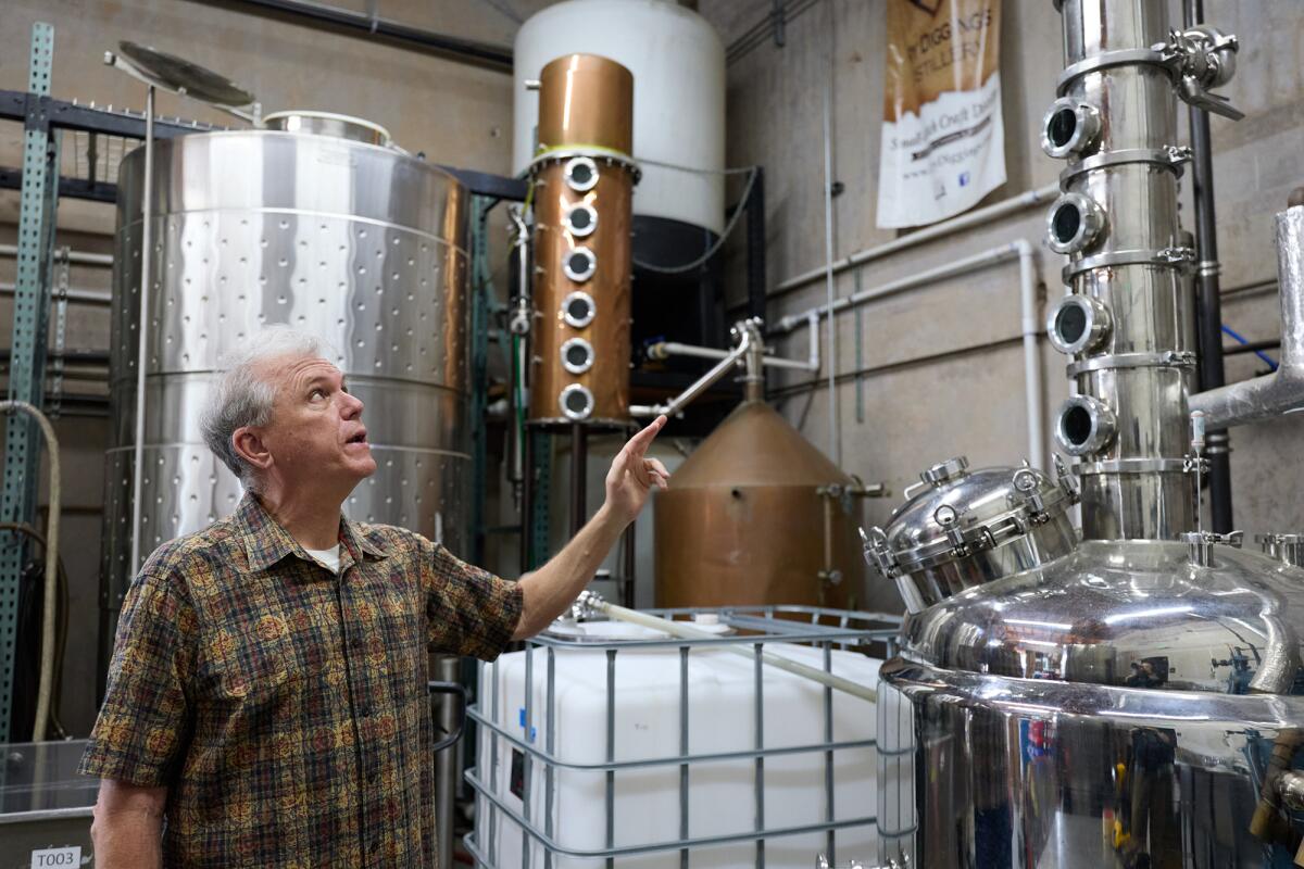 A person in a plaid shirt points upward while standing in a distillery filled with large stainless steel and copper stills.