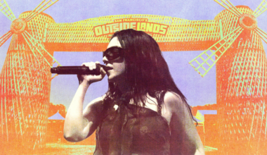 A woman with long dark hair and sunglasses sings into a microphone in front of a colorful festival entrance with two windmill structures and a sign reading "Outside Lands."