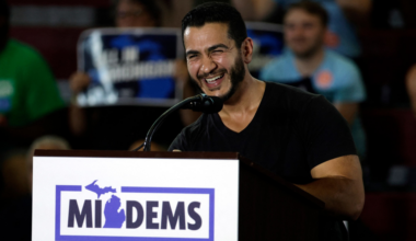 A man in a black shirt smiles and speaks at a podium with a "MI DEMS" sign in front, while an audience watches in the background.