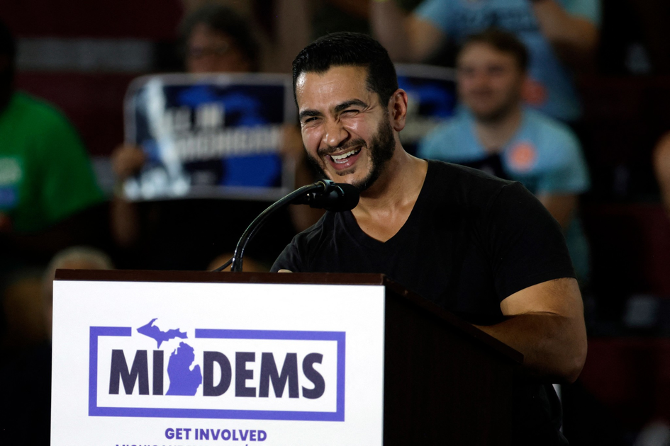 A man in a black shirt smiles and speaks at a podium with a "MI DEMS" sign in front, while an audience watches in the background.