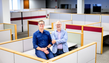 Two men, one in a blue shirt and the other in a light blue jacket, stand and sit smiling inside a mostly empty office cubicle area.