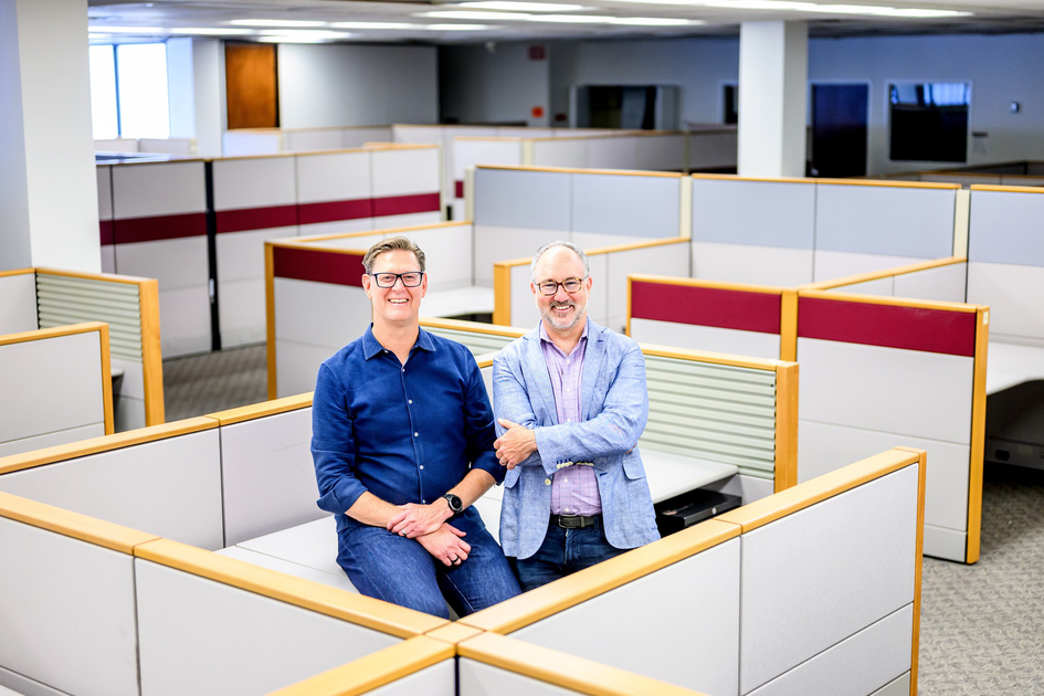 Two men, one in a blue shirt and the other in a light blue jacket, stand and sit smiling inside a mostly empty office cubicle area.