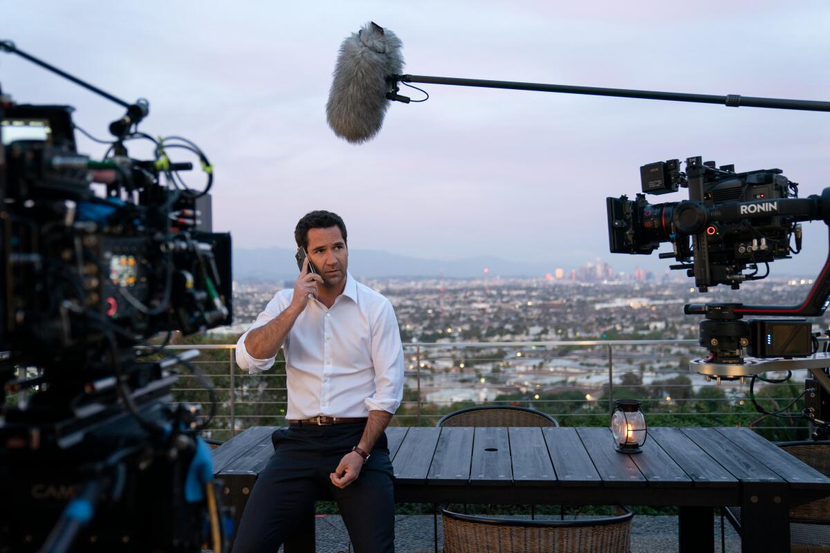 A man talks on a cellphone as a boom mic hangs over him ad two movie cameras point at him.