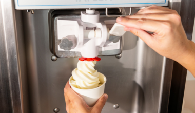 A person is dispensing soft-serve vanilla ice cream into a white paper cup from a Taylor brand machine.
