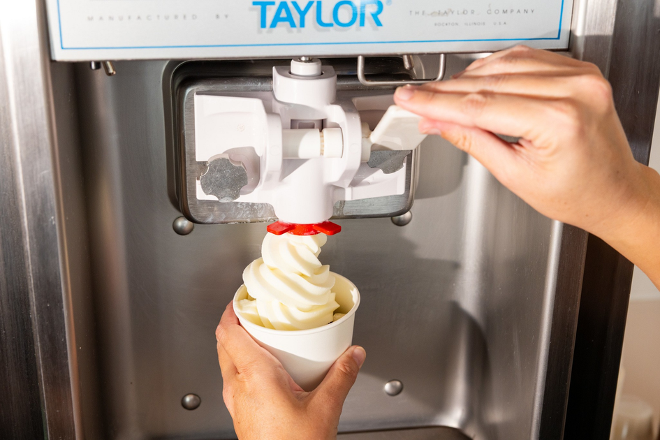 A person is dispensing soft-serve vanilla ice cream into a white paper cup from a Taylor brand machine.